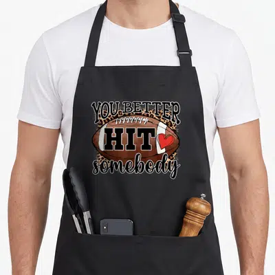 Better football apron - You Better Hit somebody text over brown football with leopard print and red heart — shown on a black cotton canvas apron against a white background