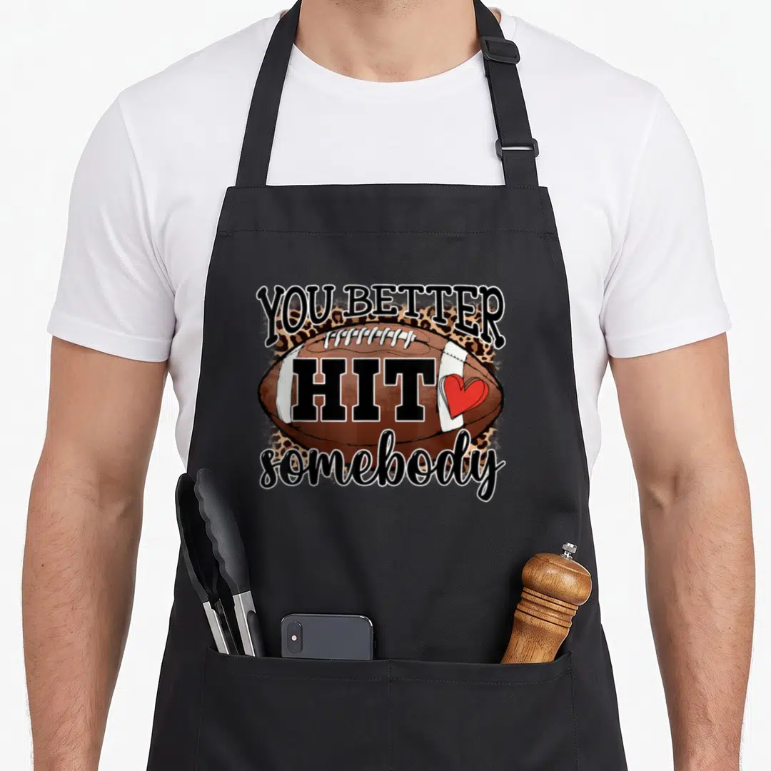 Better football apron - You Better Hit somebody text over brown football with leopard print and red heart — shown on a black cotton canvas apron against a white background