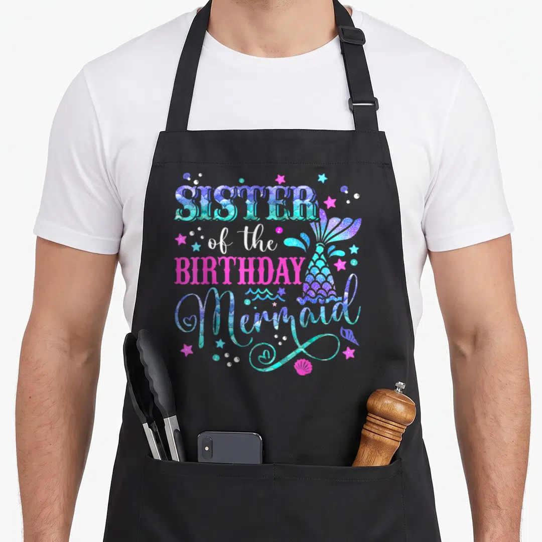 Sister birthday family aprons - Sister of the Birthday Mermaid text with colorful tail, stars, bubbles, and seashell — shown on a black cotton canvas apron against a white background