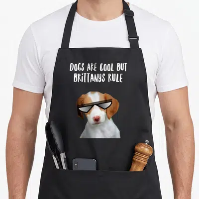 Spaniel dog rules apron - Brown and white dog wearing sunglasses below white text reading Dogs Are Cool But Brittanys — shown on a black cotton canvas apron against a white background