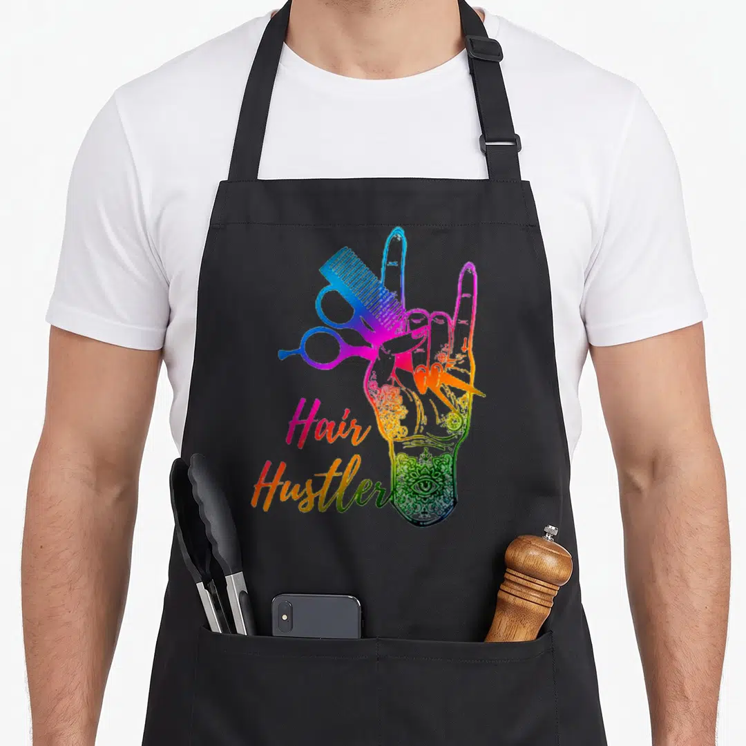 Hair tie-dye apron - Rainbow gradient hand holding scissors and comb with Hair Hustler text — shown on a black cotton canvas apron against a white background