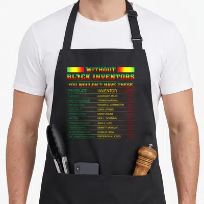 Inventor african print apron - Typography chart listing black inventors and products like traffic lights — shown on a black cotton canvas apron against a white background