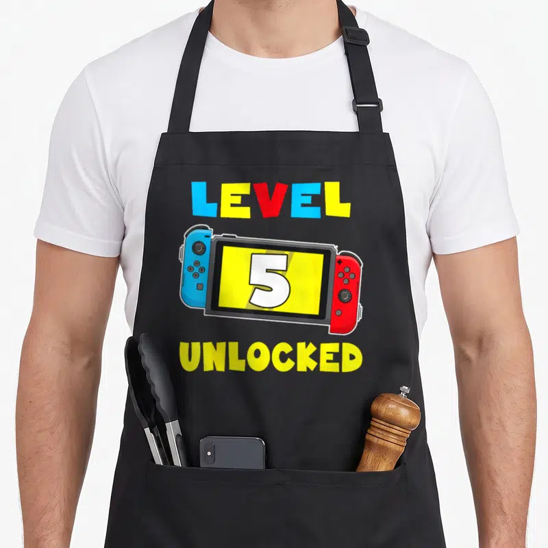 5th video game apron - Gaming console with number 5 and LEVEL 5 UNLOCKED text in yellow blue red — shown on a black cotton canvas apron against a white background