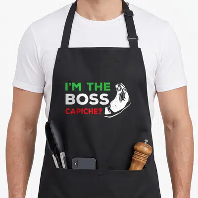 Boss italian apron - I'M THE BOSS CAPICHE? text with green white red distressed hand gesture — shown on a black cotton canvas apron against a white background
