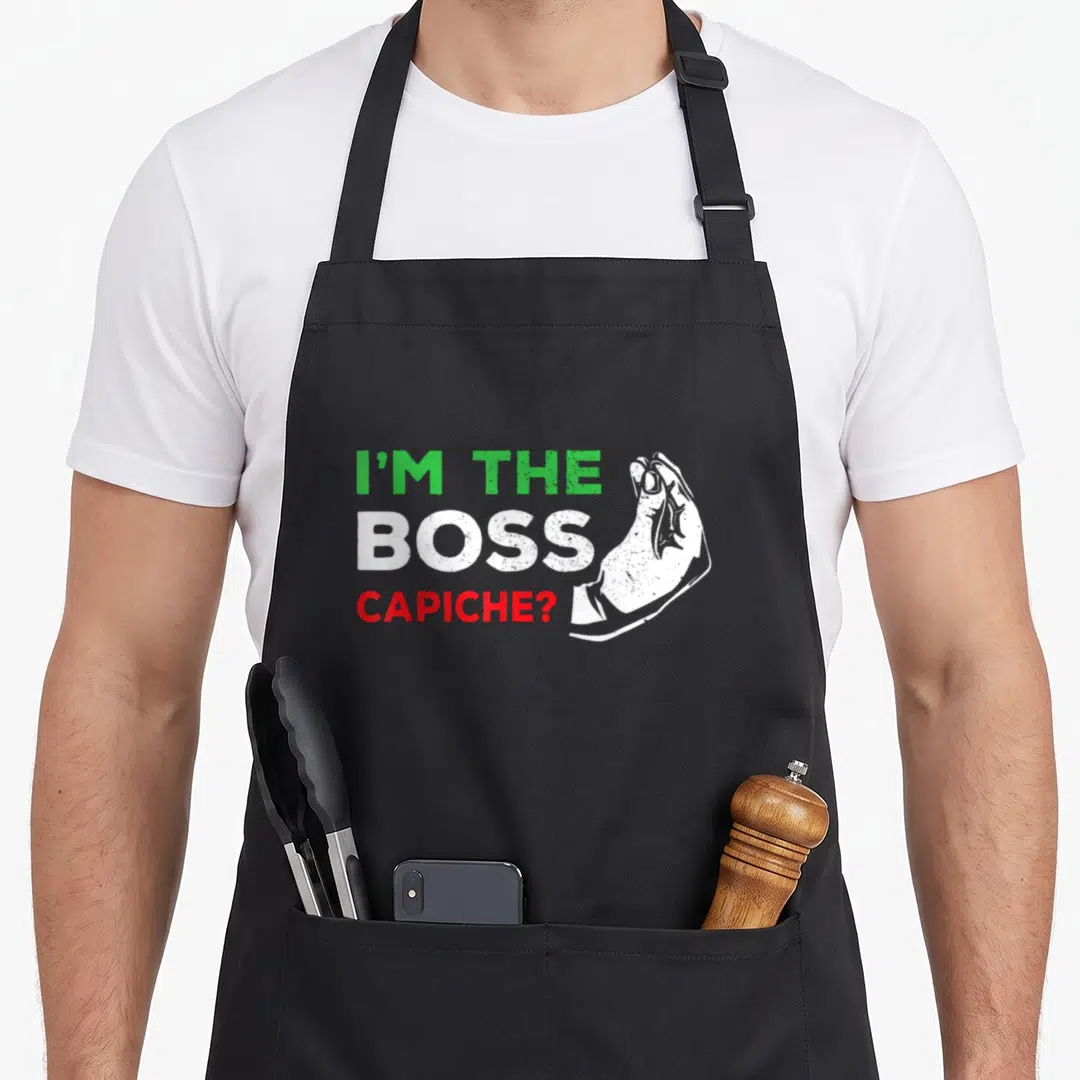 Boss italian apron - I'M THE BOSS CAPICHE? text with green white red distressed hand gesture — shown on a black cotton canvas apron against a white background