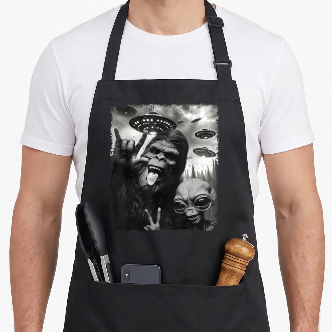 Alien bigfoot apron - Grayscale print of bigfoot making rock sign next to alien with flying ufo saucers over forest — shown on a black cotton canvas apron against a white background