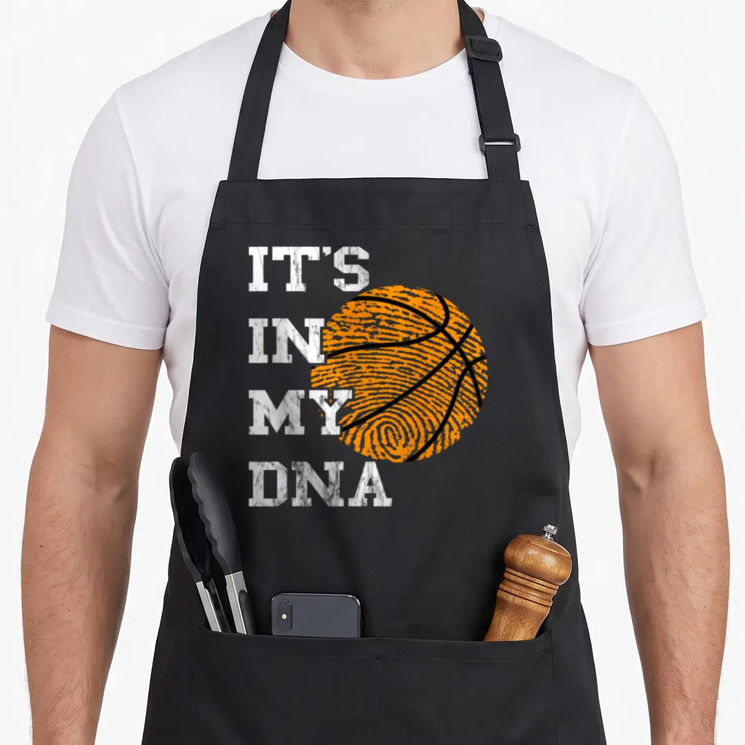 Dna basketball player apron - Stacked white text IT'S IN MY DNA next to an orange fingerprint basketball with black — shown on a black cotton canvas apron against a white background