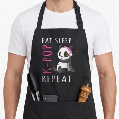 Eat kawaii apron - Cartoon panda with pink headphones surrounded by EAT SLEEP K-POP REPEAT text — shown on a black cotton canvas apron against a white background