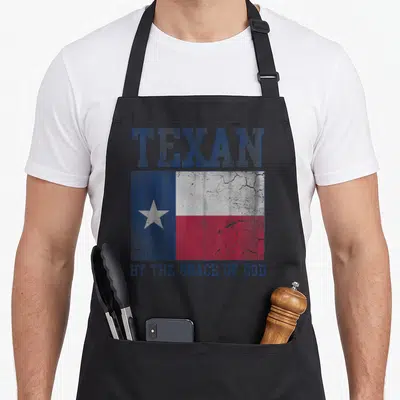 Grace texans apron - Distressed Texas flag with TEXAN text above and BY THE GRACE OF GOD below — shown on a black cotton canvas apron against a white background