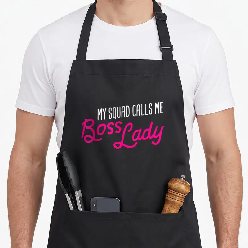 Squad boss apron - Stacked white and pink text reading MY SQUAD CALLS ME Boss Lady — shown on a black cotton canvas apron against a white background