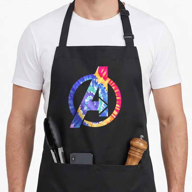 Rainbow tie dye apron - Avengers A logo filled with blue, purple, yellow, pink, and green tie-dye pattern — shown on a black cotton canvas apron against a white background