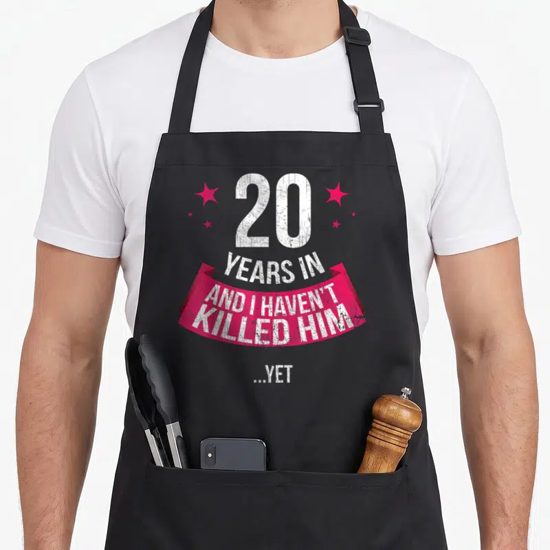 20th anniversary wedding aprons - Distressed white text 20 years in and I haven't killed him yet on pink ribbon — shown on a black cotton canvas apron against a white background