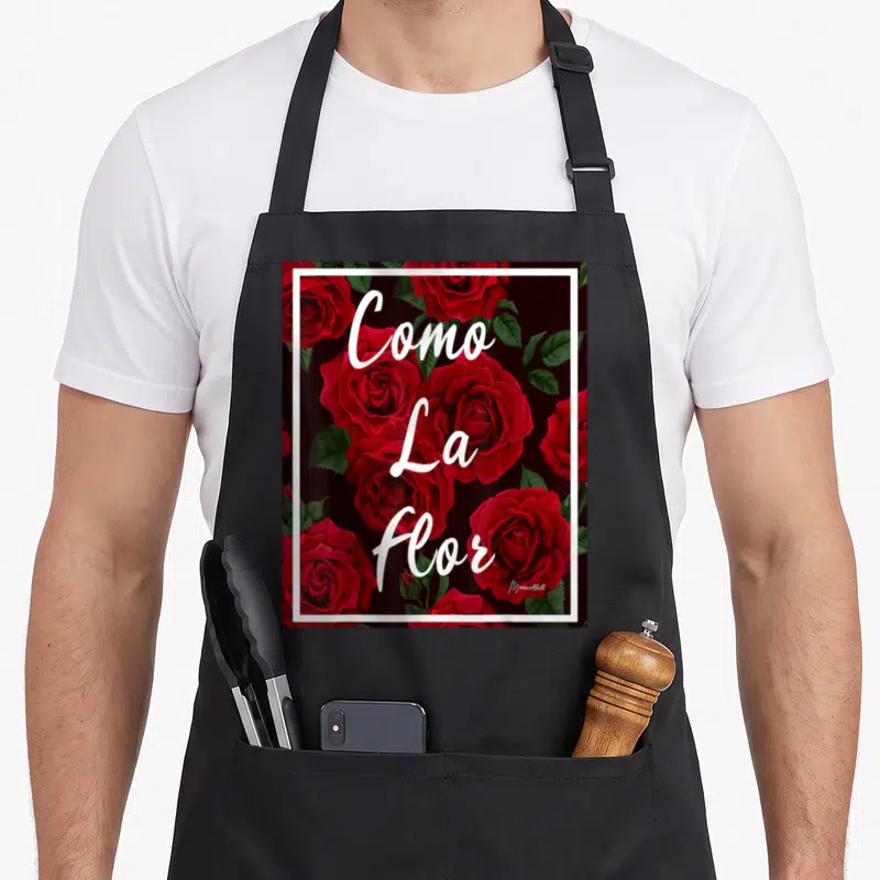 Como rose apron - Red roses and green leaves with stacked white Como La Flor text inside white border on black — shown on a black cotton canvas apron against a white background