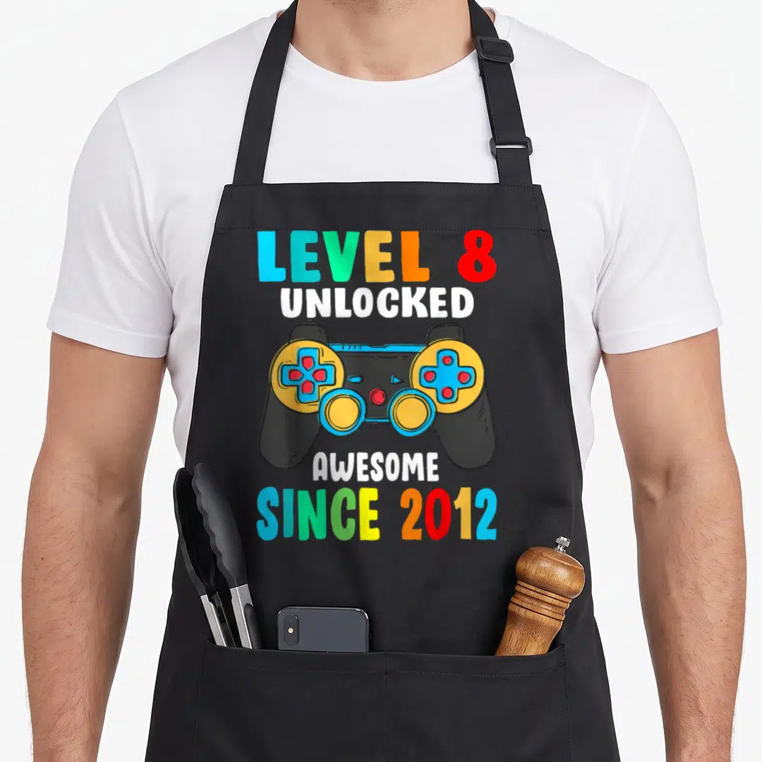 Unlocked 8th birthday apron - Cartoon black game controller with multicolored text reading LEVEL 8 UNLOCKED AWESOME — shown on a black cotton canvas apron against a white background