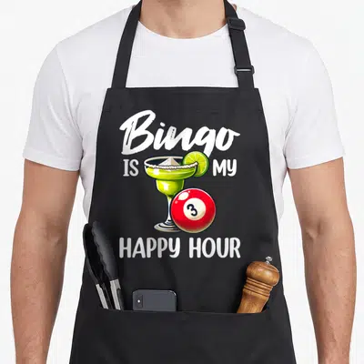 Hour bingo aprons - Cartoon green cocktail glass with lime and red number 3 bingo ball surrounded by white text — shown on a black cotton canvas apron against a white background