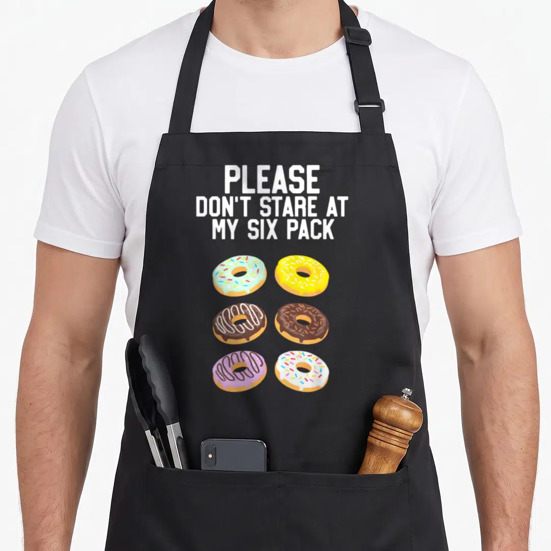 Stare donut apron - Six cartoon donuts with blue yellow brown purple and white icing under white text — shown on a black cotton canvas apron against a white background
