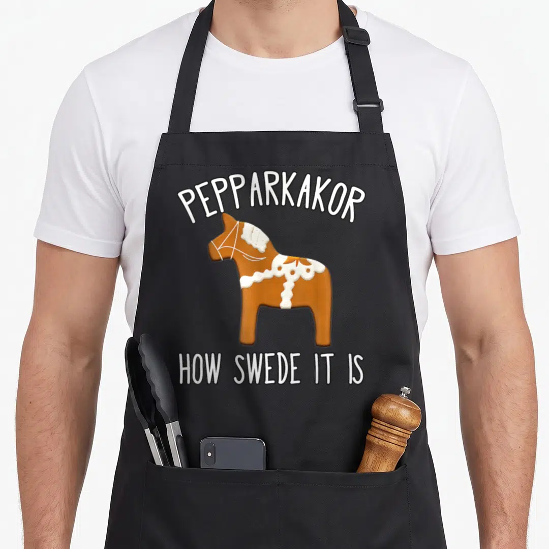 Pepparkakor women's christmas aprons - Brown Dala horse cookie with arched Pepparkakor text and straight How Swede It — shown on a black cotton canvas apron against a white background