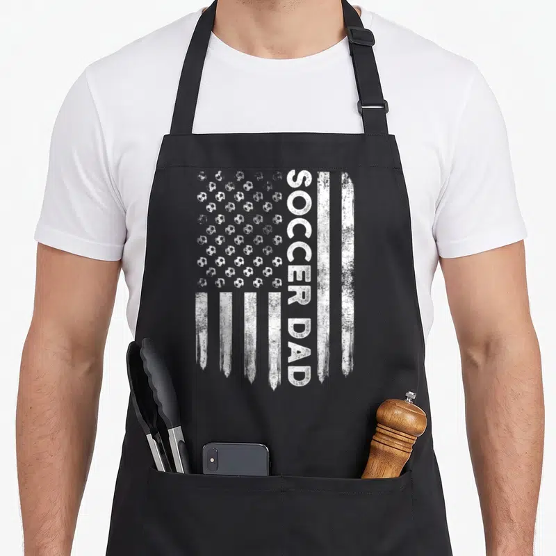 Symbol daddy apron - Distressed white flag design with soccer ball stars and vertical SOCCER DAD text — shown on a black cotton canvas apron against a white background