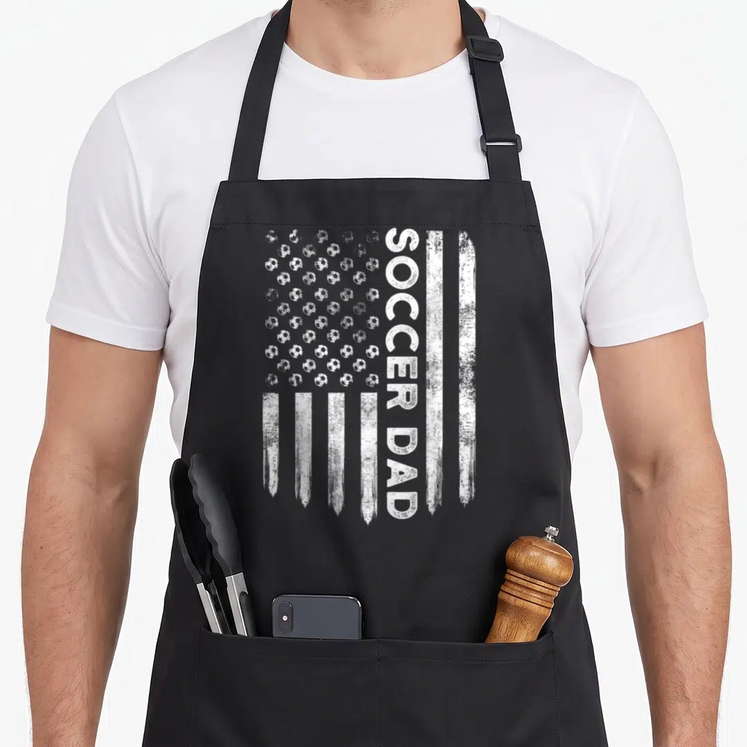 Symbol daddy apron - Distressed white flag design with soccer ball stars and vertical SOCCER DAD text — shown on a black cotton canvas apron against a white background
