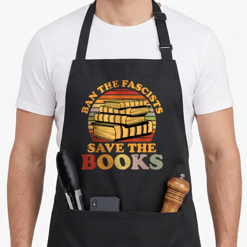 Fascist nerd apron - Retro stack of four books with striped sun background and slogan Ban The Fascists Save The Books — shown on a black cotton canvas apron against a white background