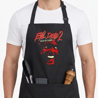 Evil bloody apron - Red monster face with EVIL DEAD 2 and DEAD BY DAWN text in distressed style on cream background — shown on a black cotton canvas apron against a white background