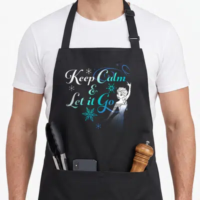Snowflake novelty apron - Keep Calm Let it Go text with Elsa and blue snowflakes in cartoon style — shown on a black cotton canvas apron against a white background