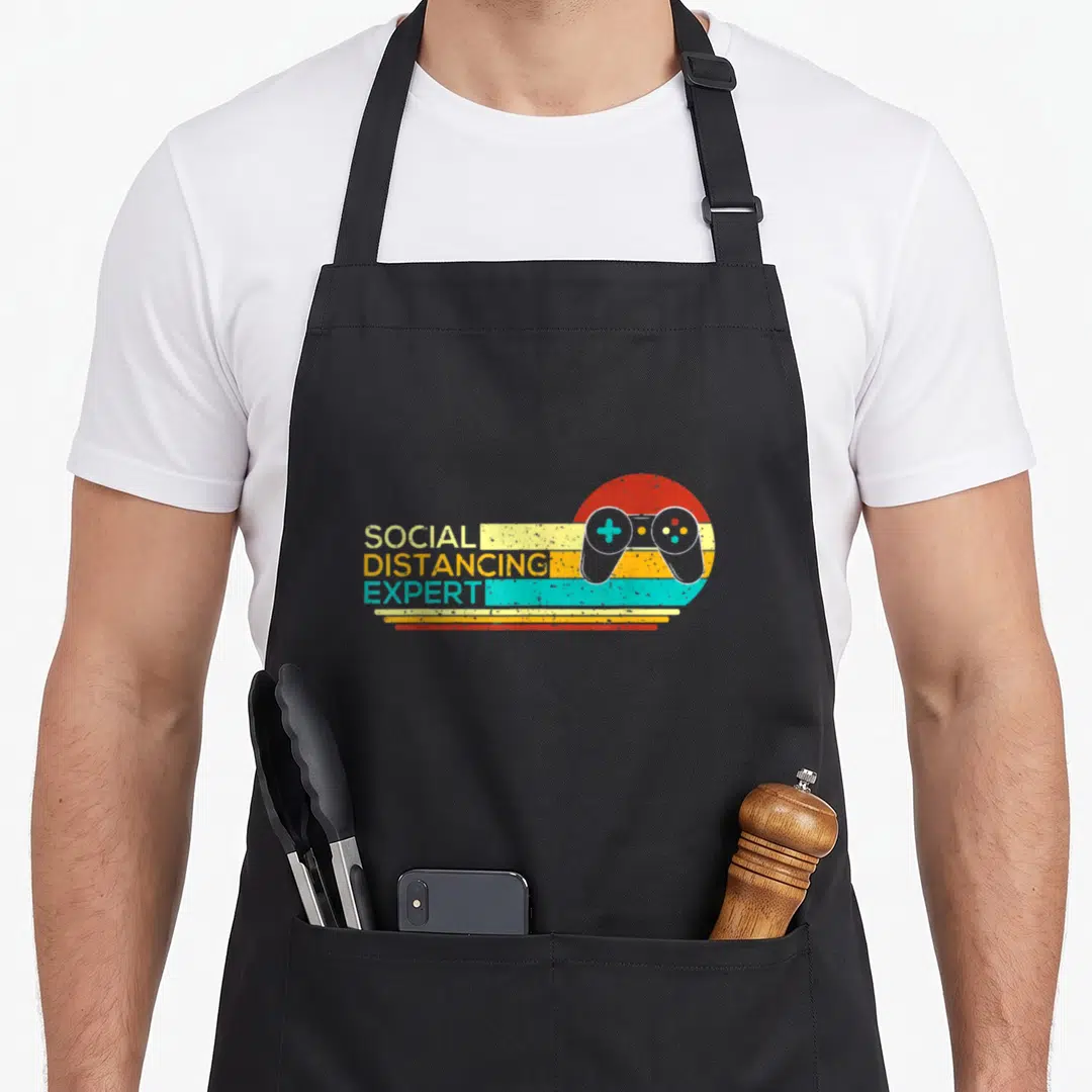 Expert gaming apron - Retro social distancing expert text with game controller and colored lines — shown on a black cotton canvas apron against a white background
