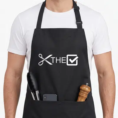 Check urban apron - White scissors, THE text, and a checked box in minimalist style — shown on a black cotton canvas apron against a white background