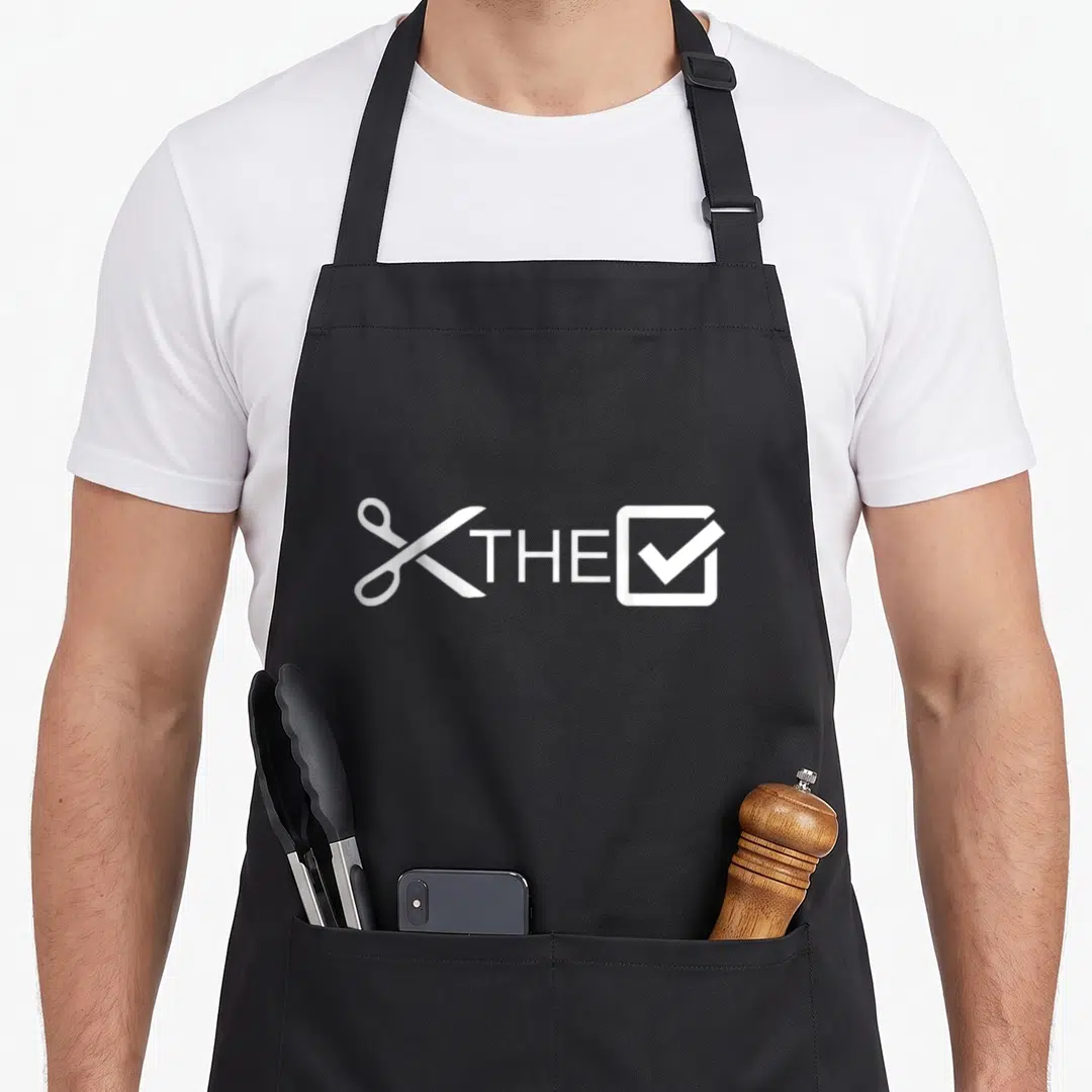 Check urban apron - White scissors, THE text, and a checked box in minimalist style — shown on a black cotton canvas apron against a white background