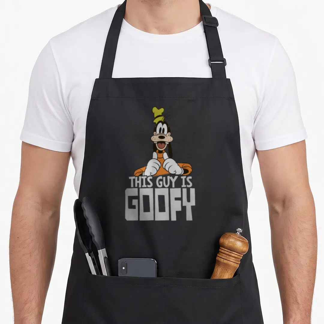 Funny worlds greatest apron - Goofy leaning over grey text THIS GUY IS GOOFY giving two thumbs up in orange and black — shown on a black cotton canvas apron against a white background
