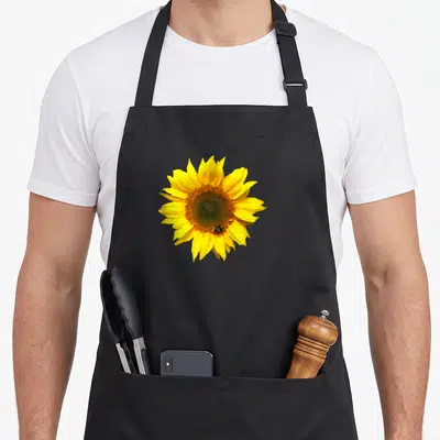 Bee floral apron - Realistic sunflower with small bee on lower petals in yellow and brown — shown on a black cotton canvas apron against a white background