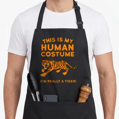 Human tiger for men women kids apron - Cartoon orange tiger with black stripes and white claws under stacked orange text — shown on a black cotton canvas apron against a white background