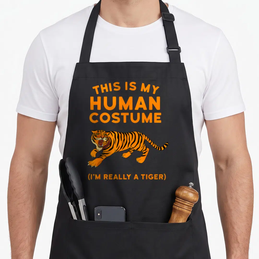 Human tiger for men women kids apron - Cartoon orange tiger with black stripes and white claws under stacked orange text — shown on a black cotton canvas apron against a white background