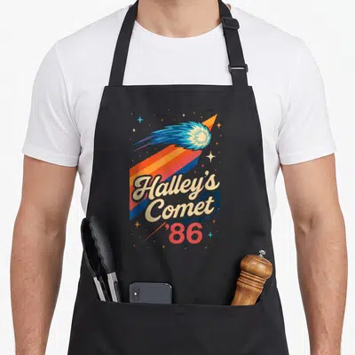 Halley comet apron - Retro 1986 comet with blue tail over diagonal orange red and blue stripes with beige text — shown on a black cotton canvas apron against a white background