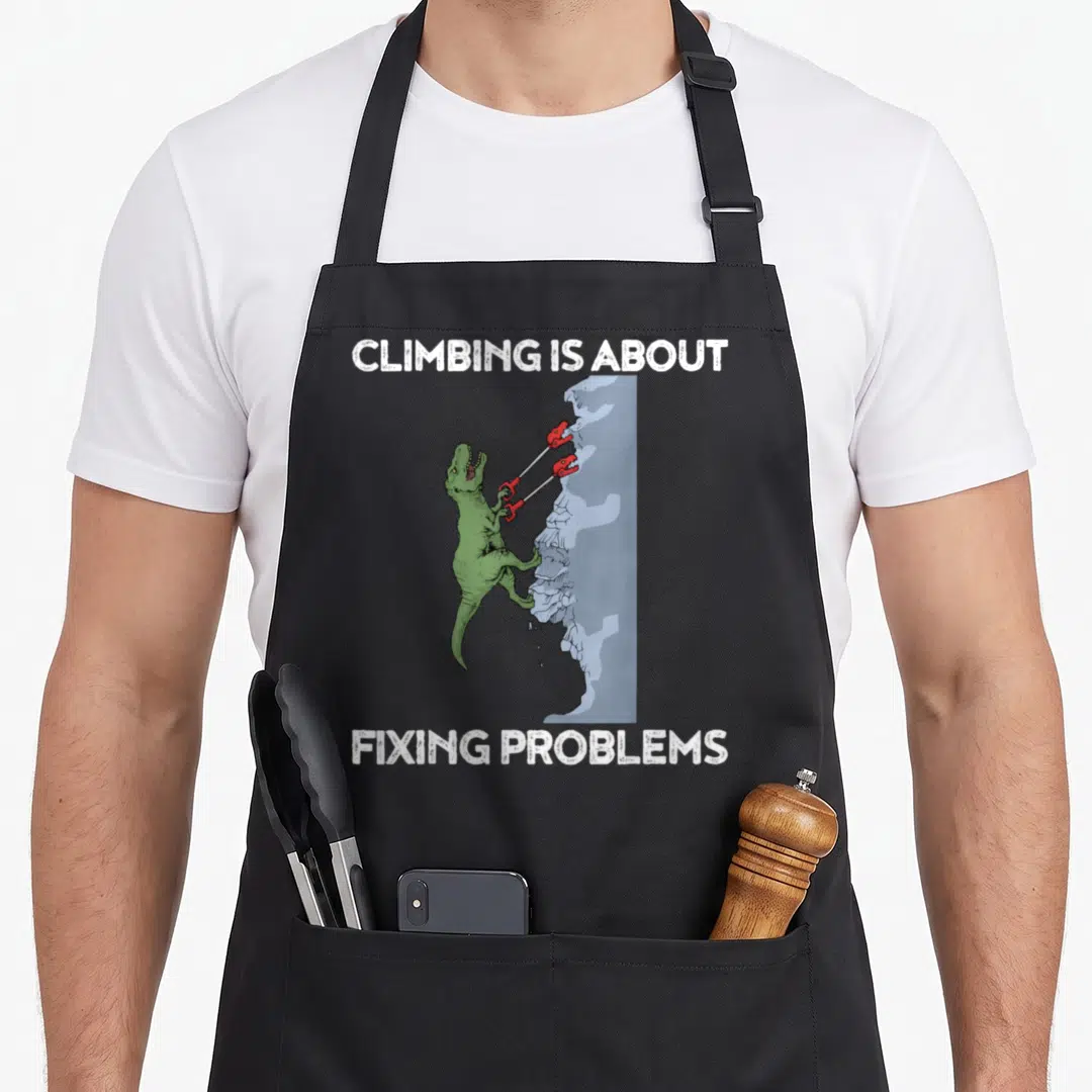 Climbing dinosaur apron - Green t-rex using red tools on blue-grey rock with white text CLIMBING IS ABOUT FIXING — shown on a black cotton canvas apron against a white background