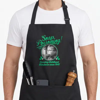 Soap christmas apron - Green script reading Soap Poisoning around grey boy portrait with holly leaves — shown on a black cotton canvas apron against a white background