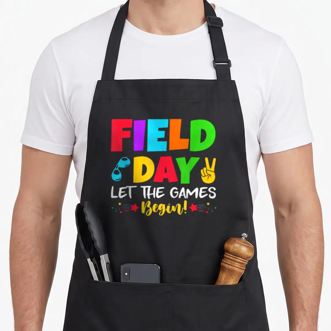 Field student teacher apron - FIELD DAY text with whistle, peace sign, and red stars in red blue yellow green white — shown on a black cotton canvas apron against a white background