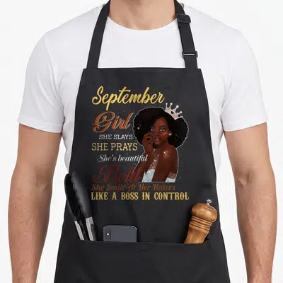 September beautiful apron - Cartoon woman with crown and stacked text reading September Girl She Slays She Prays Bold — shown on a black cotton canvas apron against a white background