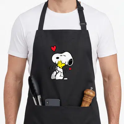 Funny lot of aprons - Snoopy hugging yellow Woodstock with one large and one small red heart in cartoon style — shown on a black cotton canvas apron against a white background