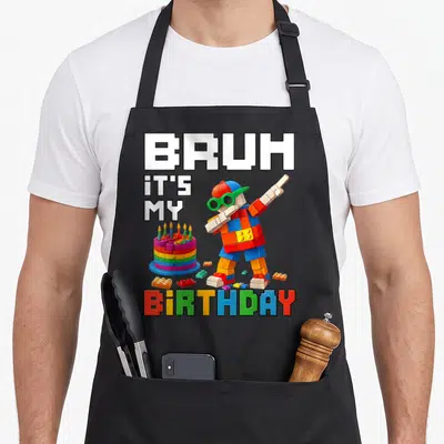 Birthday grill master apron - Lego person dabbing next to brick cake with BRUH IT'S MY BIRTHDAY text — shown on a black cotton canvas apron against a white background