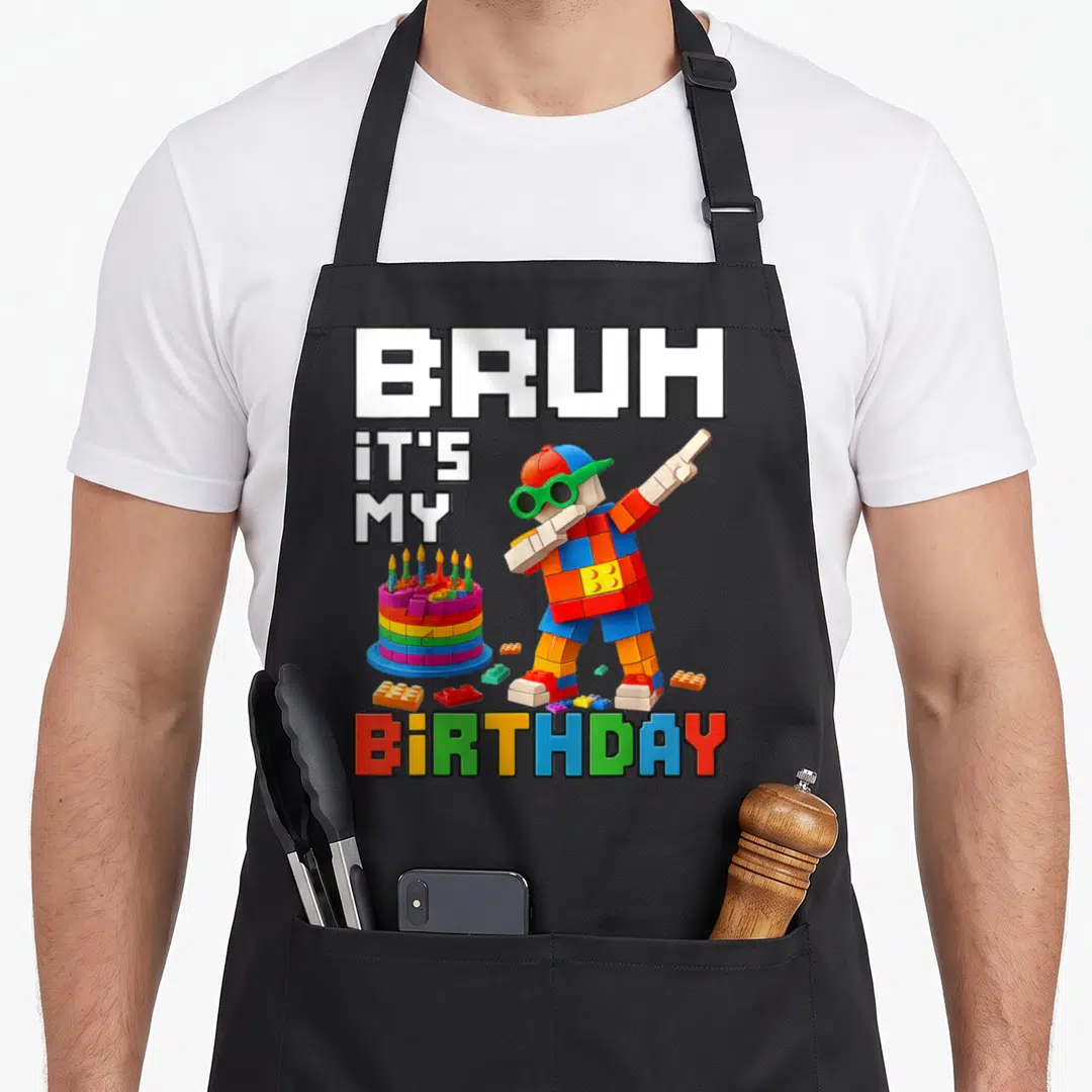 Birthday grill master apron - Lego person dabbing next to brick cake with BRUH IT'S MY BIRTHDAY text — shown on a black cotton canvas apron against a white background