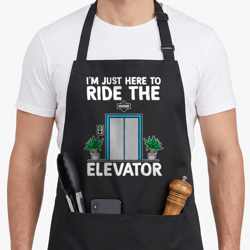 Elevator engineer apron - Cartoon blue elevator with two green potted plants and text I'M JUST HERE TO RIDE THE ELEVATOR — shown on a black cotton canvas apron against a white background