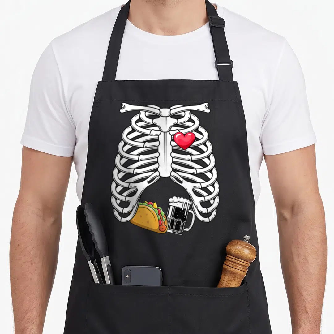 Pregnancy skeleton halloween apron - Cartoon white ribcage with red heart, taco, and beer mug in yellow and black — shown on a black cotton canvas apron against a white background