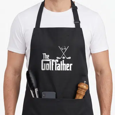 Golffather golf apron - White crossed golf clubs ball and The Golffather text in minimalist style — shown on a black cotton canvas apron against a white background