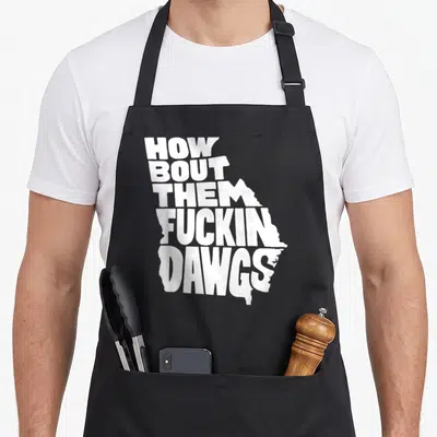 She georgia apron - White text reading How Bout Them Fuckin Dawgs shaped like the state of Georgia on black fabric — shown on a black cotton canvas apron against a white background