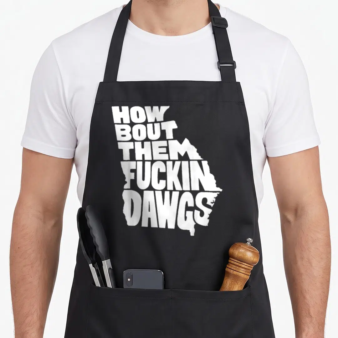 She georgia apron - White text reading How Bout Them Fuckin Dawgs shaped like the state of Georgia on black fabric — shown on a black cotton canvas apron against a white background