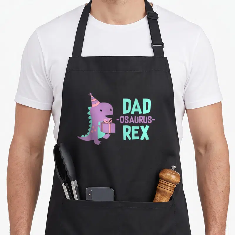 Birthday daughter family cooking aprons - Purple dinosaur with party hat holding gift next to stacked text DAD REX — shown on a black cotton canvas apron against a white background