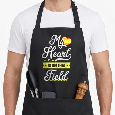 Softball field mom apron - Retro My Heart IS ON THAT Field text with yellow softball heart and three small hearts — shown on a black cotton canvas apron against a white background
