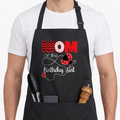 Outfit ladybug apron - MOM Of the Birthday Girl text with red ladybug and dotted line art — shown on a black cotton canvas apron against a white background