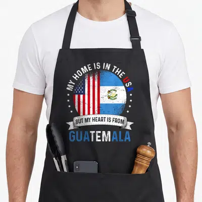 Guatemalan american flag apron - Split circle US and Guatemala flags with arched text MY HOME IS IN THE USA — shown on a black cotton canvas apron against a white background